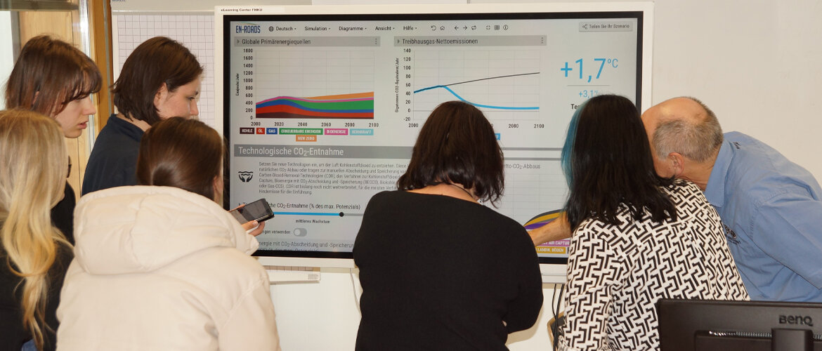 Workshop mit Klimasimulation und Datenauswertung. Teilnehmende eines Workshops betrachten gemeinsam eine Klimasimulation auf einem großen Bildschirm mit Diagrammen zu Energiequellen, Emissionen und Temperaturanstieg.