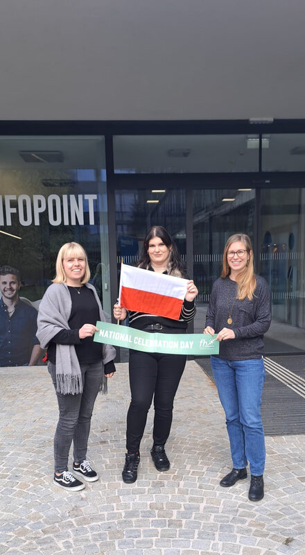 Zwei Mitarbeiterinnen mit einer Austauschstudierenden aus Polen stehen mit Schild unf Flagge vor dem FH Gebäude | © FH Kufstein Tirol