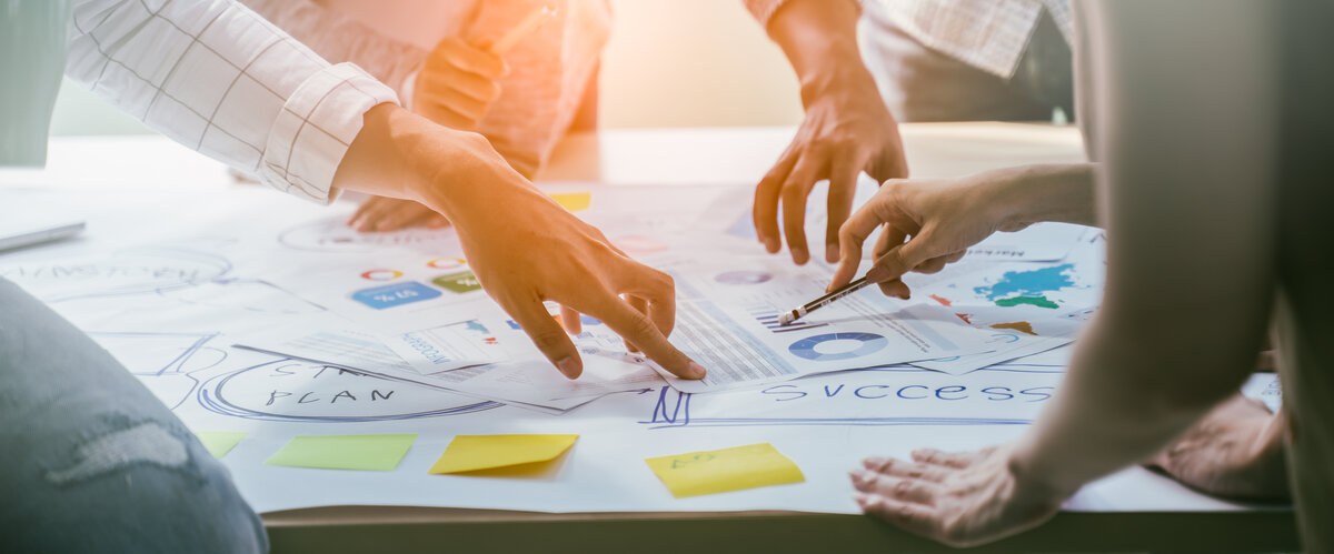 Kreatives-Meeting-mit-Post-Its-Marketing-Plan | © AdobeStock.Yingyaipumi Hände auf einem Arbeitstisch während eines Meetings, mit sichtbaren Post-Its und einem Marketing-Plan im Hintergrund. | © AdobeStock.Yingyaipumi