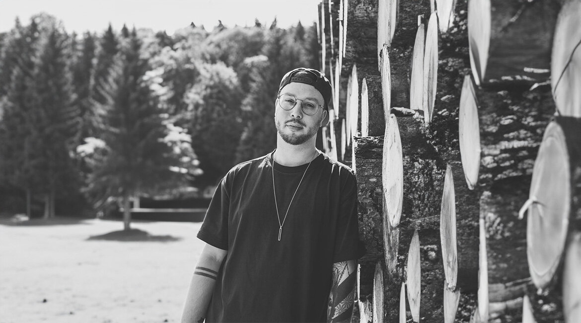 Tobias-Community-Manager-FH-Kufstein | © privat.ki-adaptiert Tobias lehnt an einem Stapel Holzstämme und blickt in die Kamera. Im Hintergrund sind Bäume und eine Wiese zu sehen. | © privat.ki-adaptiert