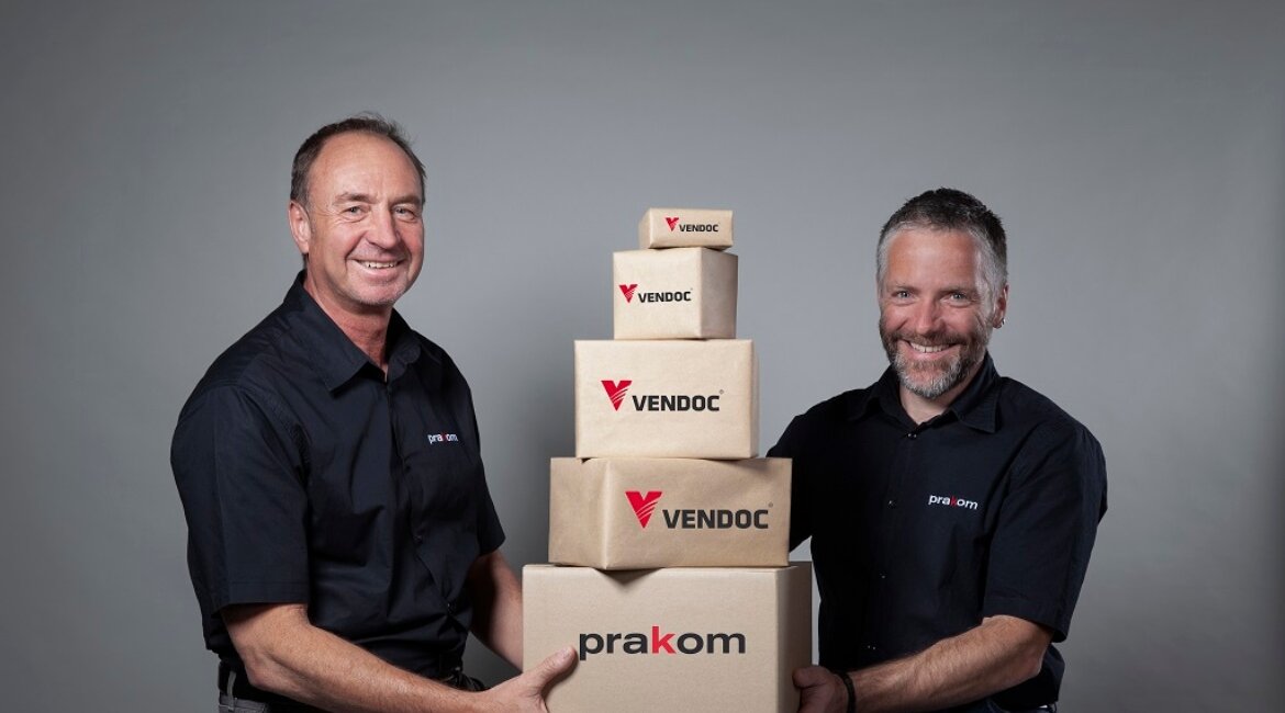 Hannes-Koidl-und-Martin-Praxmarer-präsentieren-VenDoc-Lösungen | © PraKom Zwei Männer in Prakom-Shirts halten gestapelte Kartons mit VenDoc- und Prakom-Logos vor neutralem Hintergrund. | © PraKom