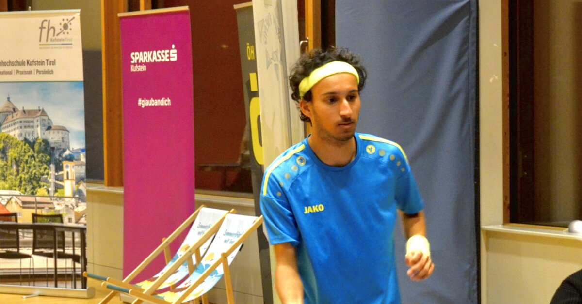 Table-Tennis-Match-FH-Kufstein | © FH Kufstein Tirol Ein Spieler in blau-gelbem Trikot mit Stirnband konzentriert sich während eines Tischtennismatches an der FH Kufstein Tirol. Im Hintergrund sind Sponsorenbanner und Liegestühle zu sehen. | © FH Kufstein Tirol