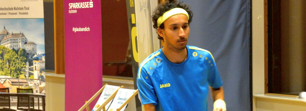 Table-Tennis-Match-FH-Kufstein | © FH Kufstein Tirol Ein Spieler in blau-gelbem Trikot mit Stirnband konzentriert sich während eines Tischtennismatches an der FH Kufstein Tirol. Im Hintergrund sind Sponsorenbanner und Liegestühle zu sehen. | © FH Kufstein Tirol