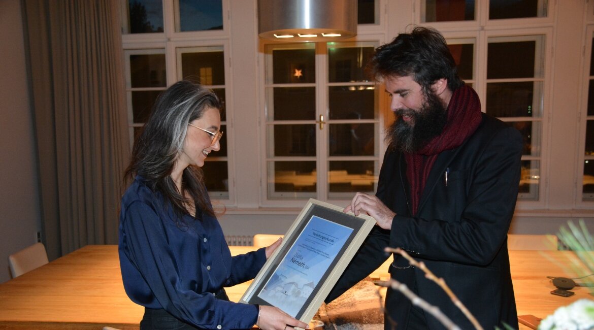 Kulturreferent Klaus Reitberger überreicht Zsófia Németh den Kufstein Thesis Award 2024. Sie hält die gerahmte Urkunde mit beiden Händen und lächelt. Im Hintergrund ist ein festlich beleuchteter Raum mit einem großen Holztisch und Fenstern zu sehen. | © Standortmarketing Kufstein