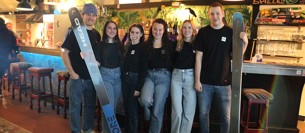 Event Team Peak & Powder Movie Night 2025 | © FH Kufstein Tirol 6 Personen stehen in Jeans und schwarzen T-Shirts vor einer Bar. Zwei haben jeweils einen Ski in der Hand. | © FH Kufstein Tirol