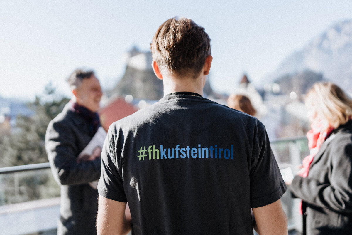 Open-House-Foto-Dachterrasse-FH-Kufstein-Tirol | © FH Kufstein Tirol Ein Student mit dem Aufdruck "#fhkufsteintirol" auf dem schwarzen T-Shirt auf der Dachterrasse der FH. Im Hintergrund sind die Umrisse der Festung zu sehen. | © FH Kufstein Tirol