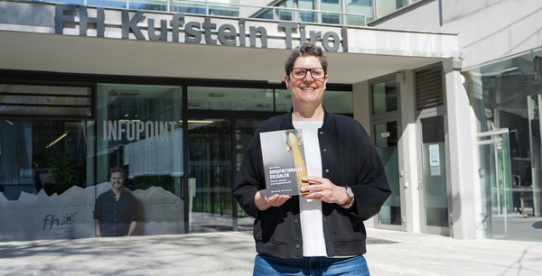 Prof. (FH) PD Dr. Agnes Bidmon vor dem Eingang der FH mit Buch in der Hand | © FH Kufstein Tirol ki-adaptiert