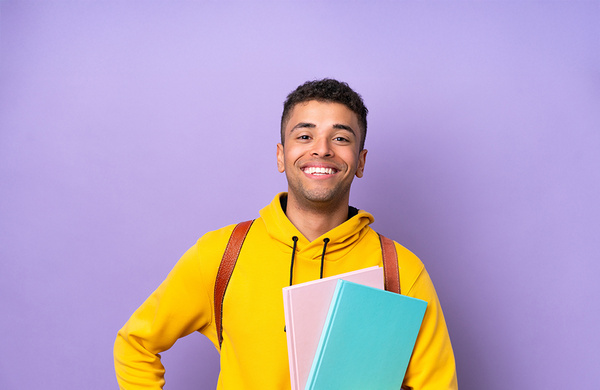 Junger Mann mit Büchern in der Hand und gelbem Pulli vor fliederfarbenen Hintergrund.  | © AdobeStock.luismolinero
