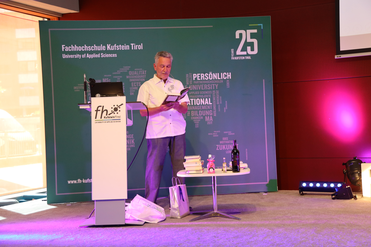 Kurt-Hoffmann-Abschiedsvorlesung-Vorlesung_Buch | © FH Kufstein Tirol Prof. (FH) Mag. Kurt Hoffmann liest auf dem Podium des Festsaals der FH Kufstein Tirol während seiner Abschiedsvorlesung aus einem Buch. | © FH Kufstein Tirol