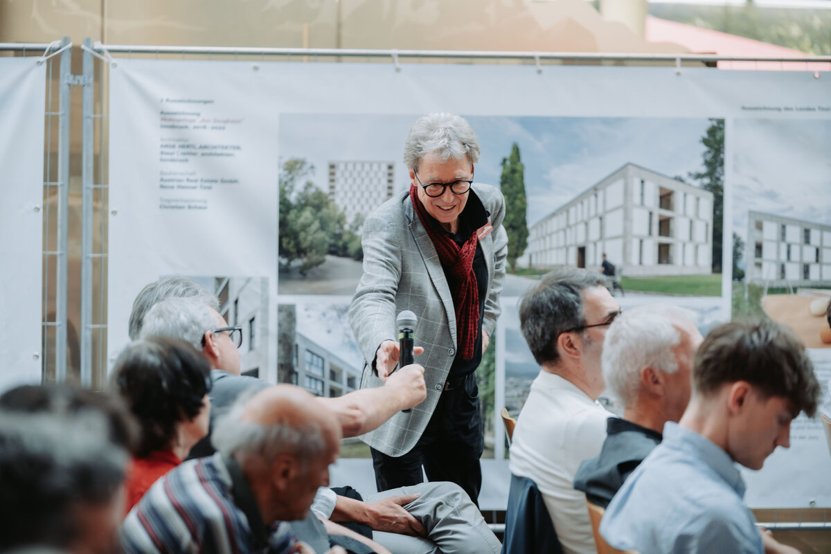 ausstellung-neues-bauen-ramsauer | © Fotoschmiede Ein Mann reicht einem anderen Mann ein Mikrofon. Him Hintergrund ein Bauzaunbanner. | © Fotoschmiede