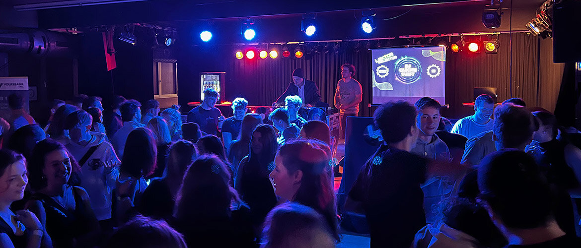 Party-Stimmung bei Lektor Selektor 2023 an der FH Kufstein Tirol. Volles Haus bei der Veranstaltung Lektor Selektor 2023: Ein DJ legt auf der Bühne auf, während zahlreiche Studierende im bunt beleuchteten Club tanzen und feiern.