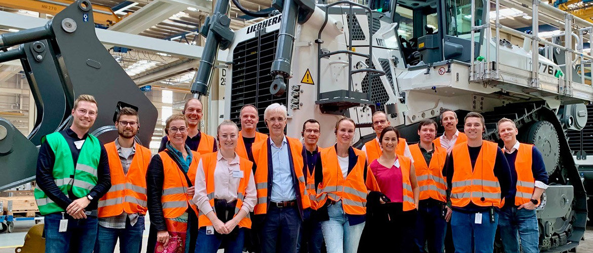 Gruppe von Teilnehmer:innen in orangenen Sicherheitswesten bei einer Werksbesichtigung von Liebherr in Telfs vor einer großen Baumaschine.