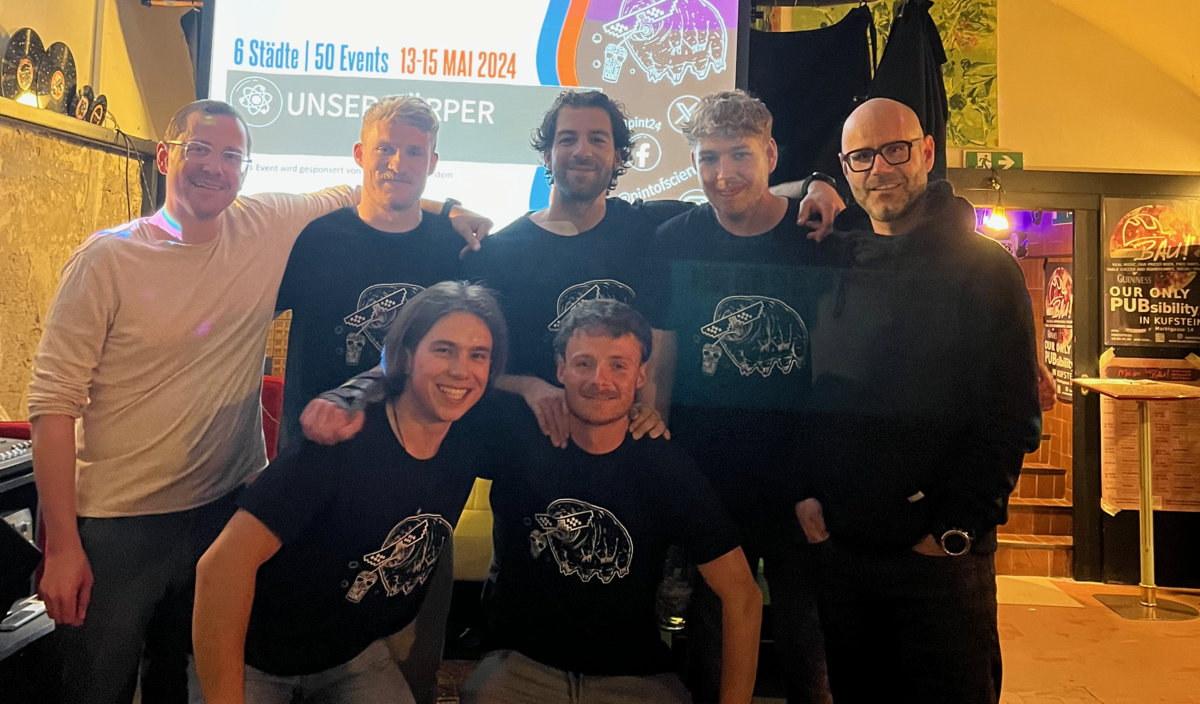 pint-of-sciences-festival-team-kufsteiner-eventlocation-bau Organisationsteam des Pint of Sciences Festivals in der Kufsteiner Eventlocation BAU posiert vor einer Präsentationsleinwand.