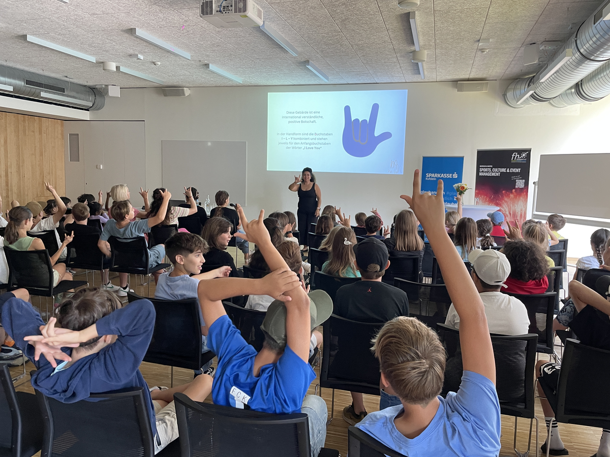 Eine Gruppe von Schüler:innen macht in einem Vorlesungsraum der FH Kufstein Tirol die Handzeichen der gehörlosen Pädagogin nach.