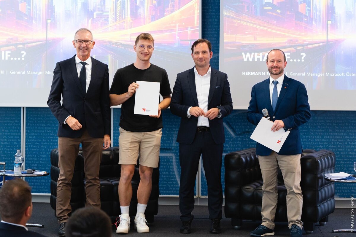 Eine Gruppe von drei Männern, einer davon jung und in kurzen Hosen mit einer Urkunde in der Hand, auf einer Bühne.