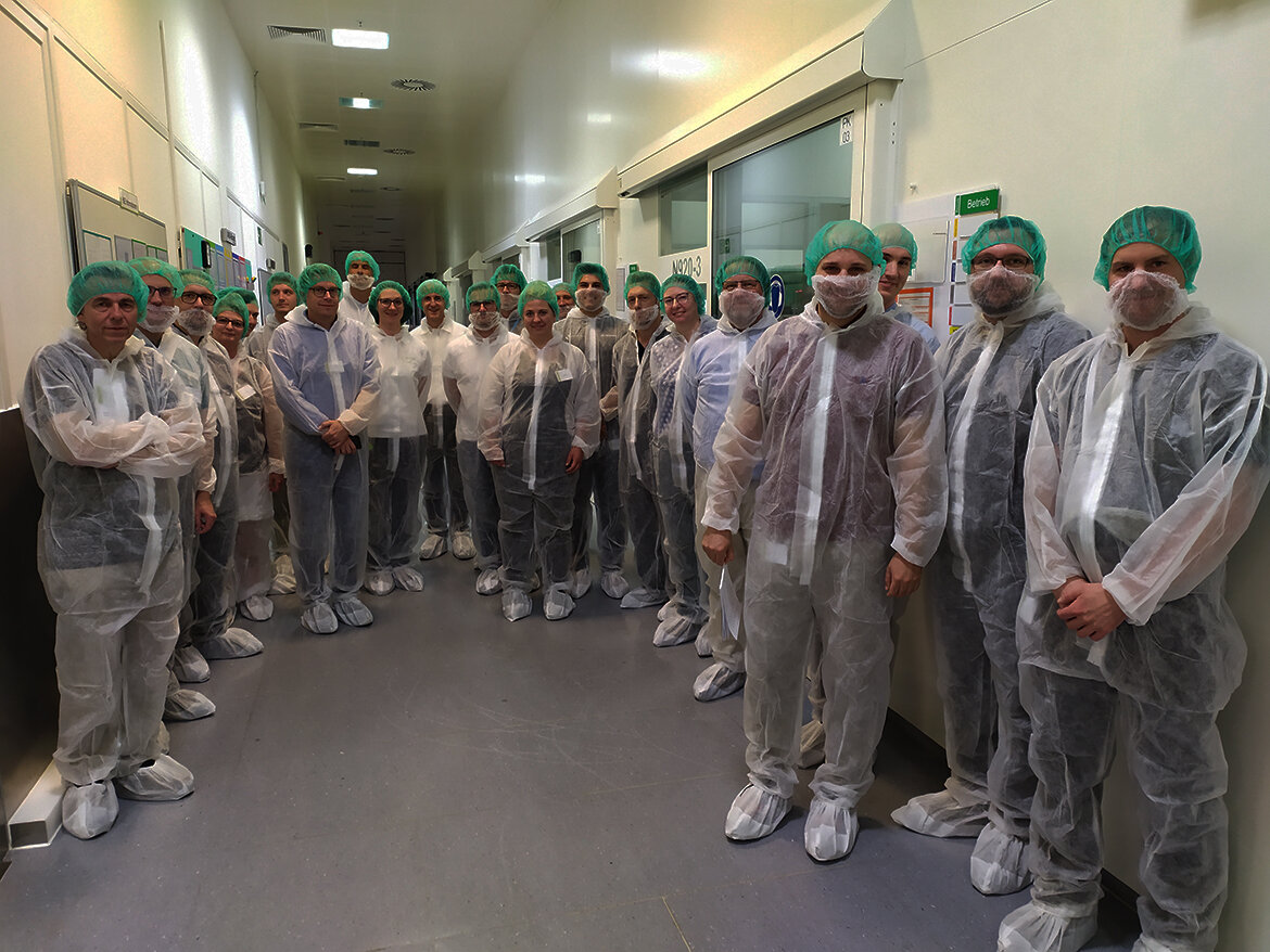 Gruppe von Personen in steriler Schutzkleidung und Hauben, die sich in einem hell beleuchteten Fertigungsgang befinden.