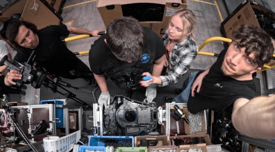 Studierende der FH Kufstein Tirol und ein Kameraprofi des eLearning Centers richten gemeinsam mit einem Produktionsmitarbeiter von STIHL Tirol die Kamera für Prozessaufnahmen an einer Mähroboter-Montagelinie ein.