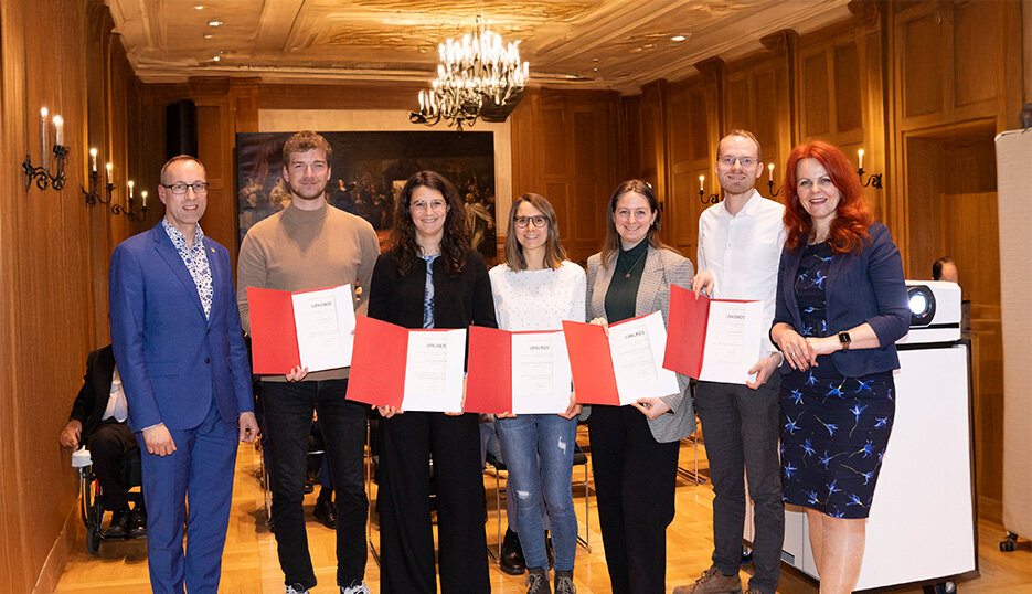 Gruppenfoto in einem festlichen Raum: FH-Rektor Prof. (FH) Dr. Mario Döller, Preisträger:innen Quentin Hirtl, Franziska Boroviczeny, Lea Carnuth, Selina-Maria Schiller, Lukas Hartleif und Landesrätin Cornelia Hagele halten Urkunden.