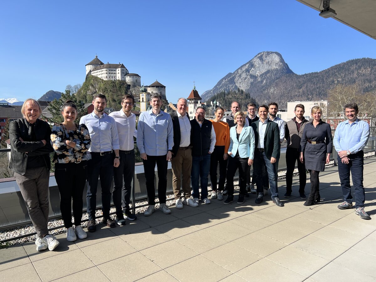 Eine Gruppe Menschen steht auf einer Terrasse, im Hintergrund blauer Himmel und Berge