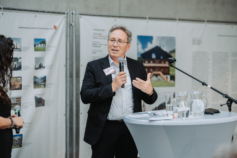 Ein Mann mit einem Mikrofon steht an einem Stehtisch vor einem Bauzaunbanner.