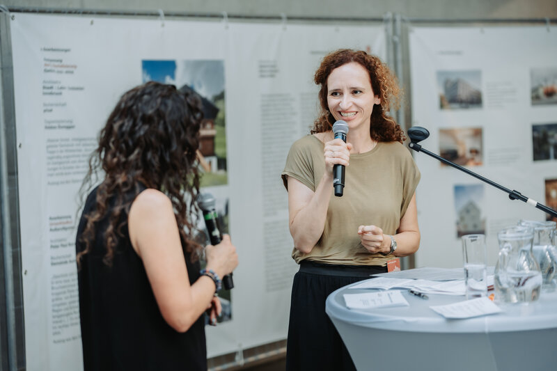 Eine Frau mit Mikrofon steht vor einem Bauzaunbanner.