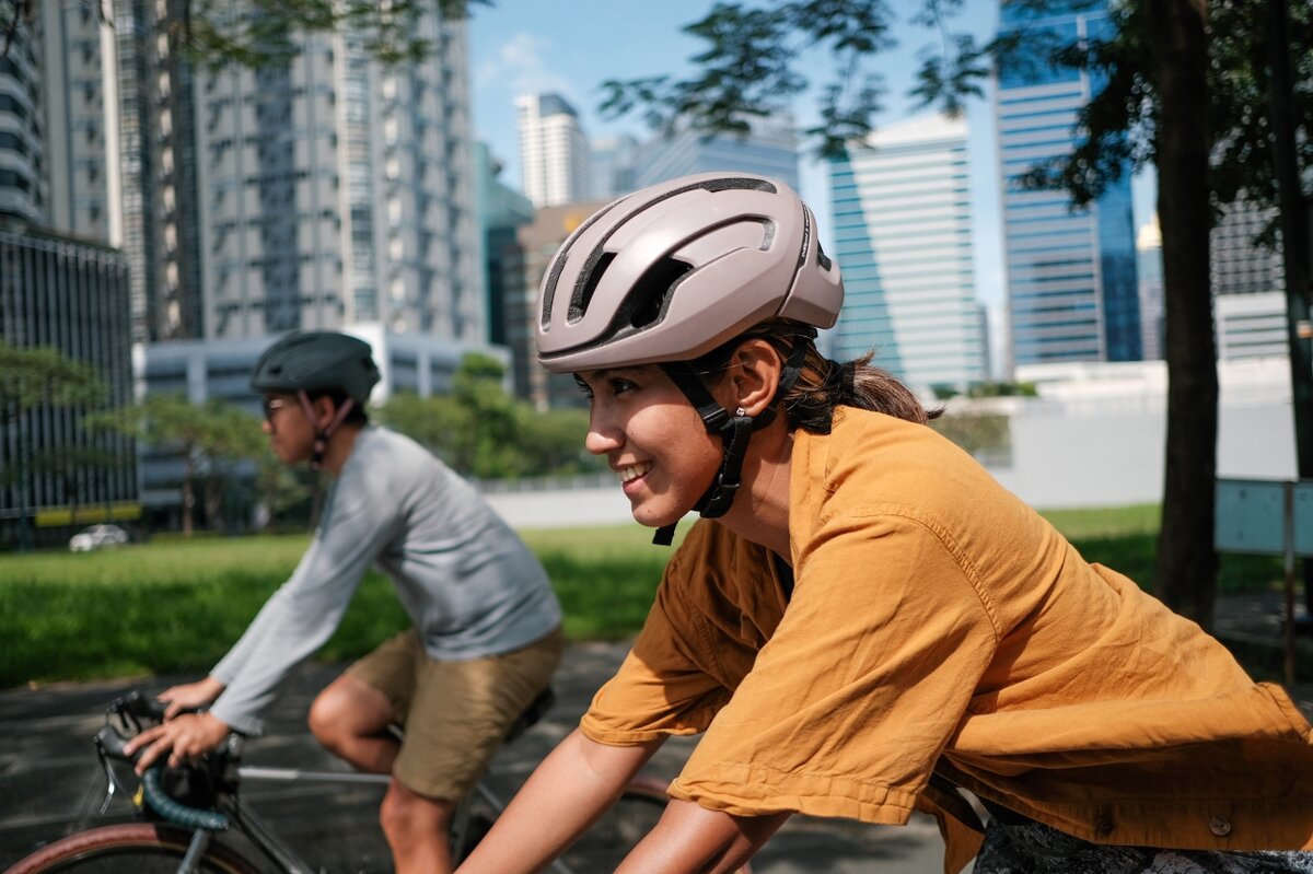 Zwei Radfahrer:innen mit Fahrradhelm fahren durch ein städtisches Gebiet mit modernen Hochhäusern im Hintergrund.