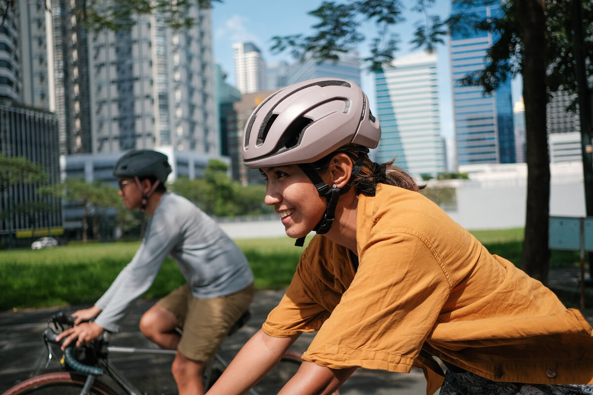 Zwei Radfahrer:innen mit Fahrradhelm fahren durch ein städtisches Gebiet mit modernen Hochhäusern im Hintergrund.