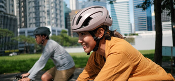 Zwei Radfahrer:innen mit Fahrradhelm fahren durch ein städtisches Gebiet mit modernen Hochhäusern im Hintergrund.