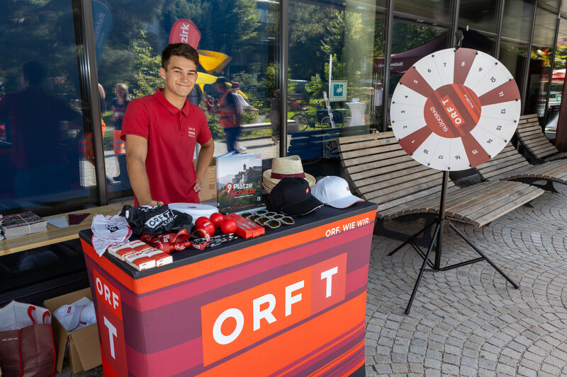 Junger Mann am ORF-Stand bei der Sommerfrische 2025 mit Glücksrad und Merchandise-Artikeln.