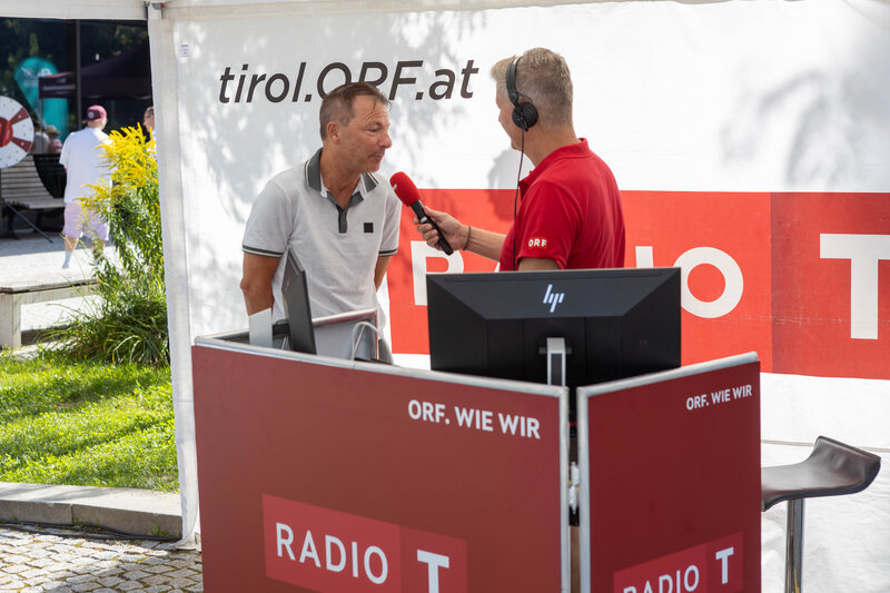 ORF-Moderator im roten Poloshirt interviewt Thomas Madritsch, Geschäftsführer der FH Kufstein Tirol, bei der Sommerfrische 2025 am Radiostand.