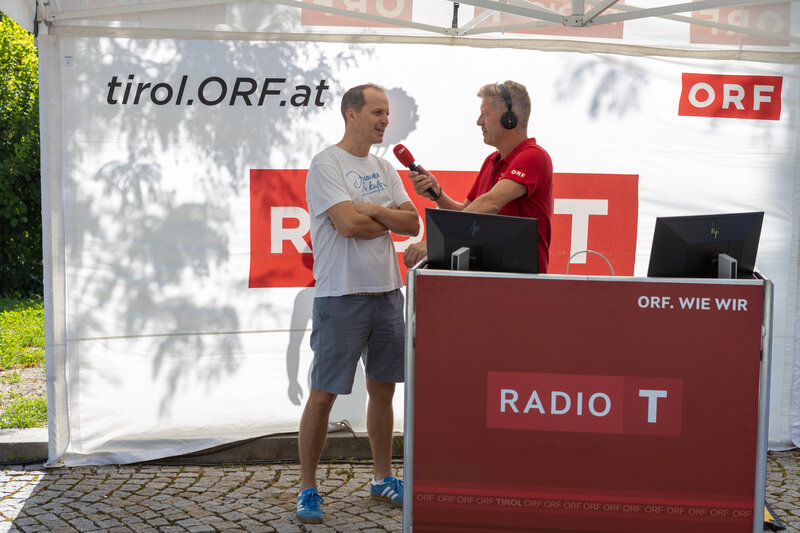 ORF-Moderator im roten Poloshirt interviewt einen Mann im weißen T-Shirt mit der Aufschrift „Discover Kufstein“ bei der Sommerfrische 2025 am Radiostand.