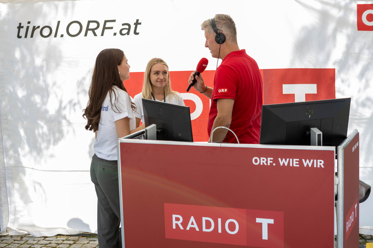 ORF-Moderator im roten Poloshirt interviewt zwei Studentinnen im weißen T-Shirt mit der Aufschrift „FH Kufstein Tirol“ am Radiostand der Sommerfrische 2025.
