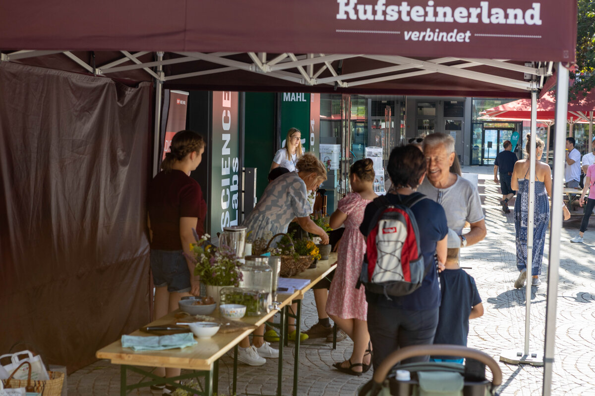 Besucherinnen und Besucher nehmen an einem Workshop unter dem Pavillon „Kufsteinerland verbindet“ teil. Auf einem langen Tisch sind Pflanzen, Kräuter und Materialien für kreative Arbeiten ausgestellt.