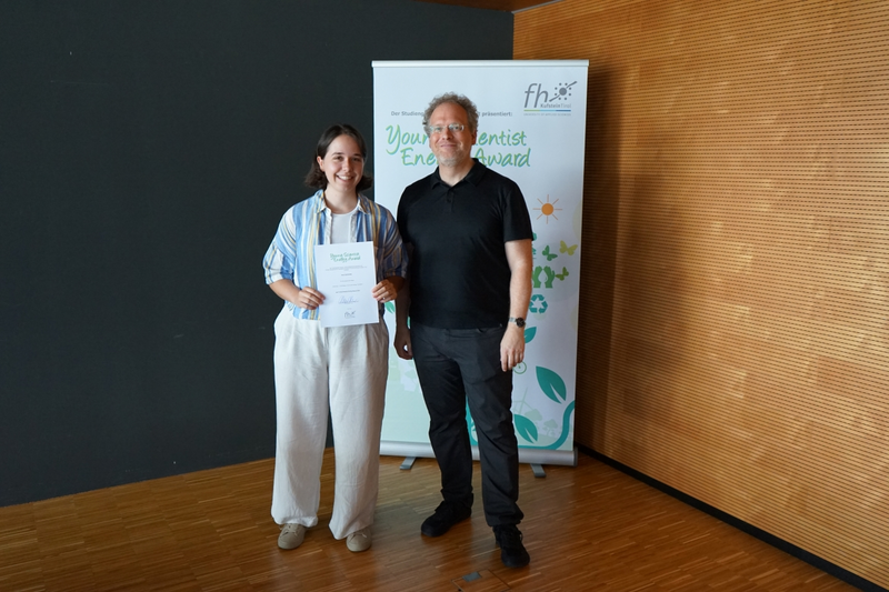 Teilnehmerin des Young Scientist Energy Award mit Urkunde und Juror vor Banner.