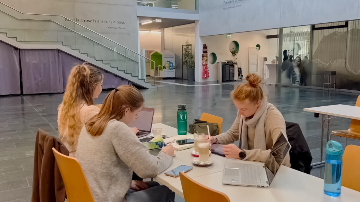 Zwei Studierende und eine Lehrende arbeiten mit Laptops und Unterlagen in der Aula der FH Kufstein Tirol.