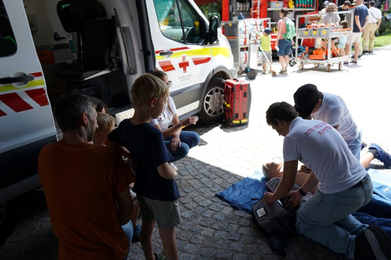 Sanitäter führen erste Hilfe an einer Puppe vor und Kinder schauen an der Seite zu.