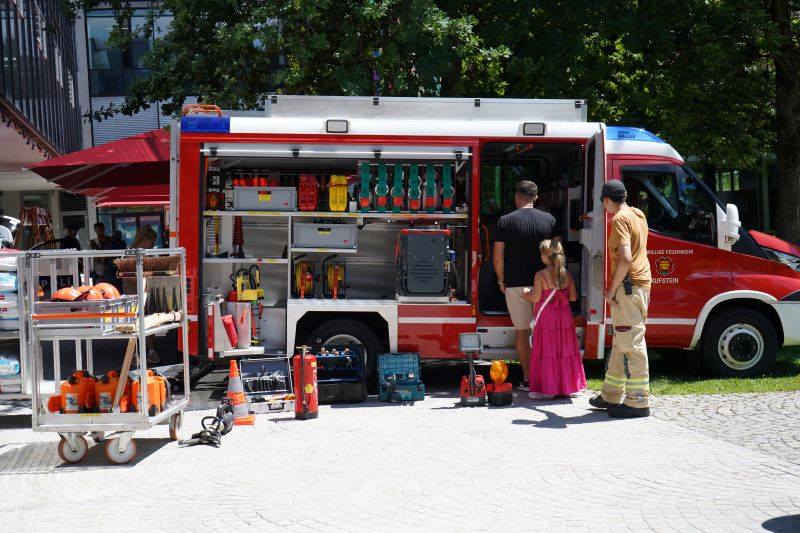 Ein Feuerwehrmann, ein Mann und ein Kind stehen vor einem Feuerwehrauto im Stadtpark vor dem Gebäude der FH Kufstein Tirol.