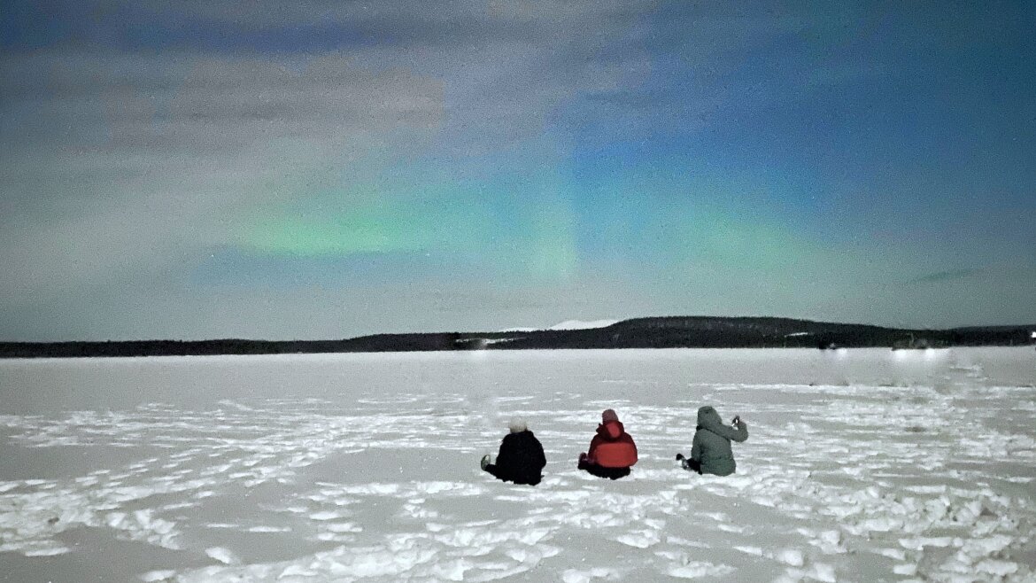 Drei Studierende sitzen auf einer weiten flachen Schneefläche und beobachten die eindrucksvollen grünen Schlieren der Nordlichter am Himmel.