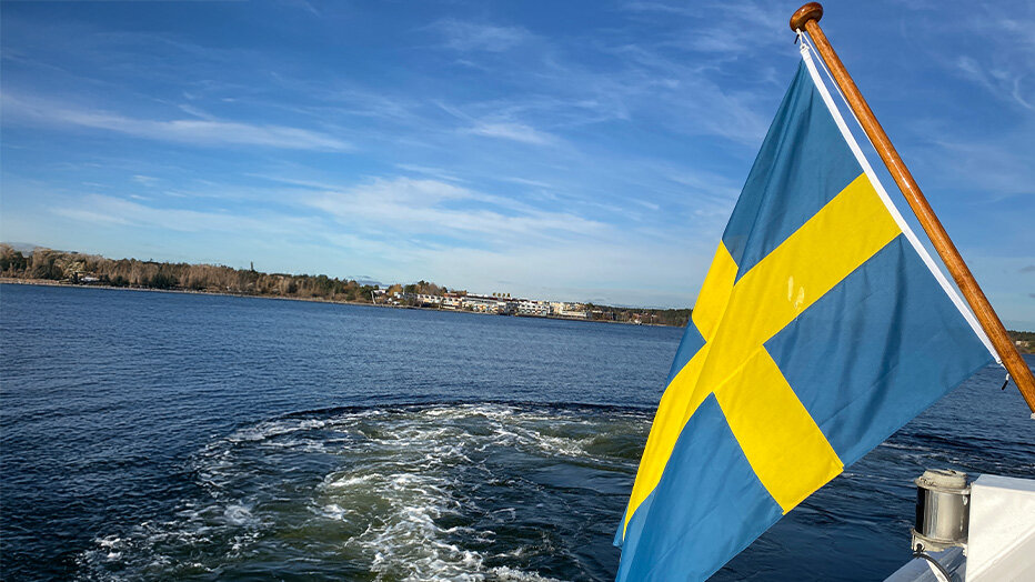 Der Blick vom Heck eines Bootes an dem eine Schwedische Flagge hängt, auf das durch den Antrieb verwirbelte Wasser, mit einer kleinen Stadt in der Ferne.
