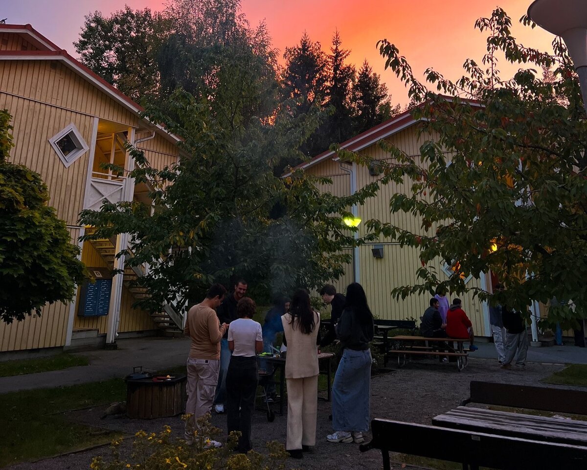 Einige Studierende stehen im Innenhof zwischen zwei gelben Häusern zusammen im Kreis um einen Grill herum und unterhalten sich unter orange gefärbtem Himmel.