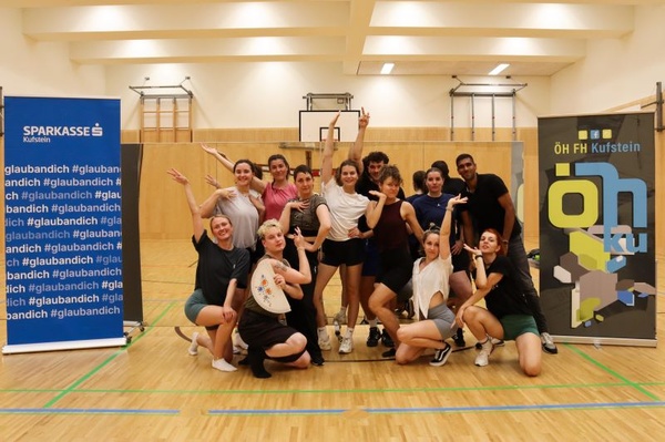 Eine Gruppe von Studierenden posiert in einer Turnhalle nach einem Tanzworkshop des Praxisprojekts Q & U, zwischen Bannern der Sparkasse Kufstein und der ÖH FH Kufstein Tirol. | © FH Kufstein Tirol