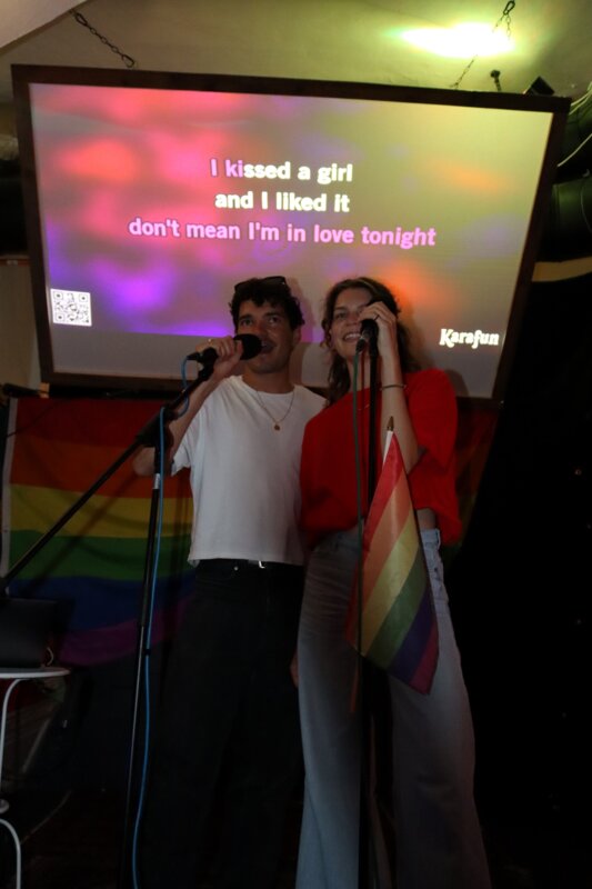 Zwei Studierende singen bei der Karaoke Night des Praxisprojekts QYOU an der FH Kufstein Tirol. Sie stehen auf einer Bühne vor einer Leinwand mit Liedtext und halten Mikrofone, während eine Regenbogenflagge sichtbar ist.