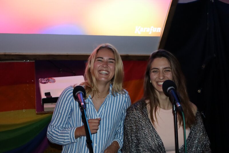 Zwei Studierende stehen lachend auf der Bühne der Karaoke Night des Praxisprojekts QYOU an der FH Kufstein Tirol. Beide halten Mikrofone in der Hand, im Hintergrund ist eine Leinwand und eine Regenbogenflagge zu sehen.