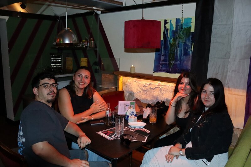 Vier Studierende sitzen an einem Tisch in einer gemütlichen Bar und nehmen am Pub Quiz „Drag Your Brain Out“ im Rahmen des Praxisprojekts QYOU der FH Kufstein Tirol teil. Getränke und Quiz-Unterlagen liegen auf dem Tisch.