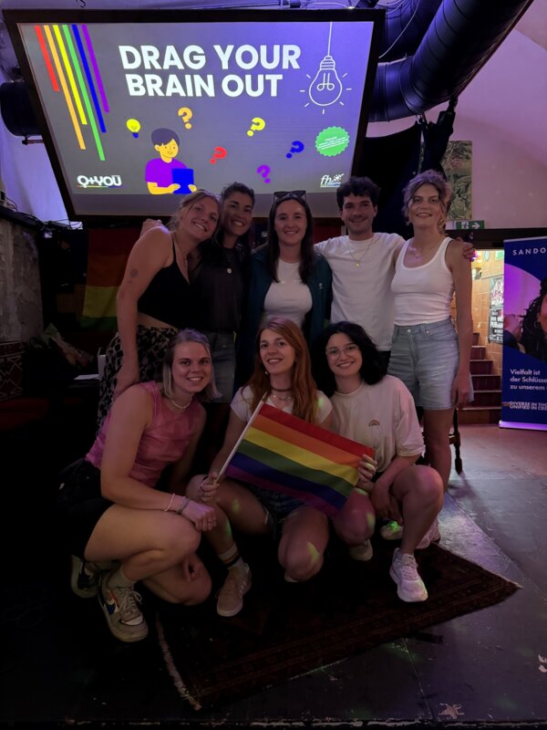 Eine Gruppe von acht Studierenden posiert lächelnd vor der Leinwand mit der Aufschrift „Drag Your Brain Out“ beim Pub Quiz des Praxisprojekts QYOU an der FH Kufstein Tirol. Eine Person hält eine Regenbogenflagge, im Hintergrund sind Regenbogenfarben und Symbole des Quizdesigns zu sehen.
