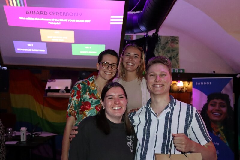 Vier Studierende des Gewinnerteams posieren lachend bei der Award Ceremony des Pub Quiz „Drag Your Brain Out“ im Rahmen des Praxisprojekts QYOU an der FH Kufstein Tirol. Im Hintergrund ist die Leinwand mit der Ankündigung der Siegerehrung zu sehen.