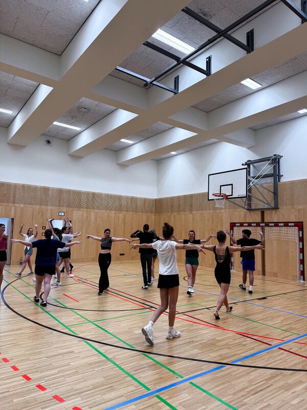 Mehrere Studierende nehmen in einer Turnhalle am QYOU-Tanzworkshop teil. Sie stehen in einem Kreis und bewegen sich synchron mit ausgebreiteten Armen, während die Trainerin die Übung anleitet.