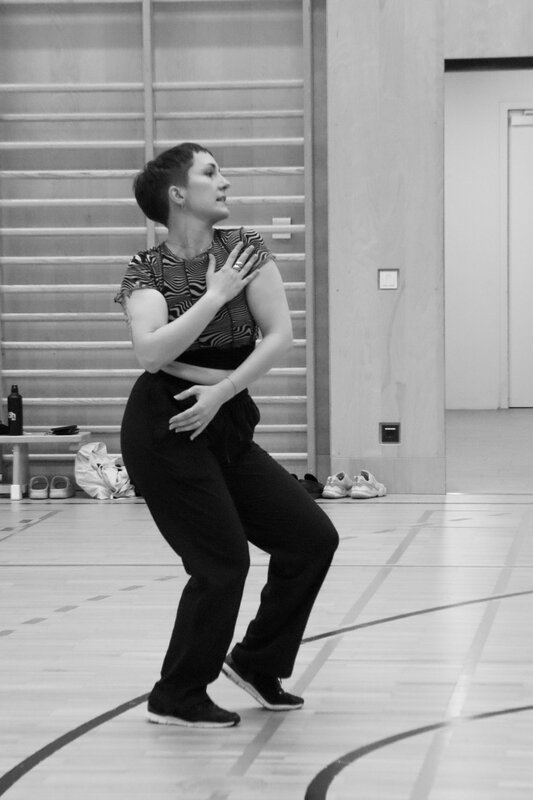 Eine Teilnehmerin des QYOU-Tanzworkshops steht in einer Turnhalle und tanzt mit einer ausdrucksstarken Geste. Das Bild ist in Schwarz-Weiß aufgenommen und betont den künstlerischen Ausdru