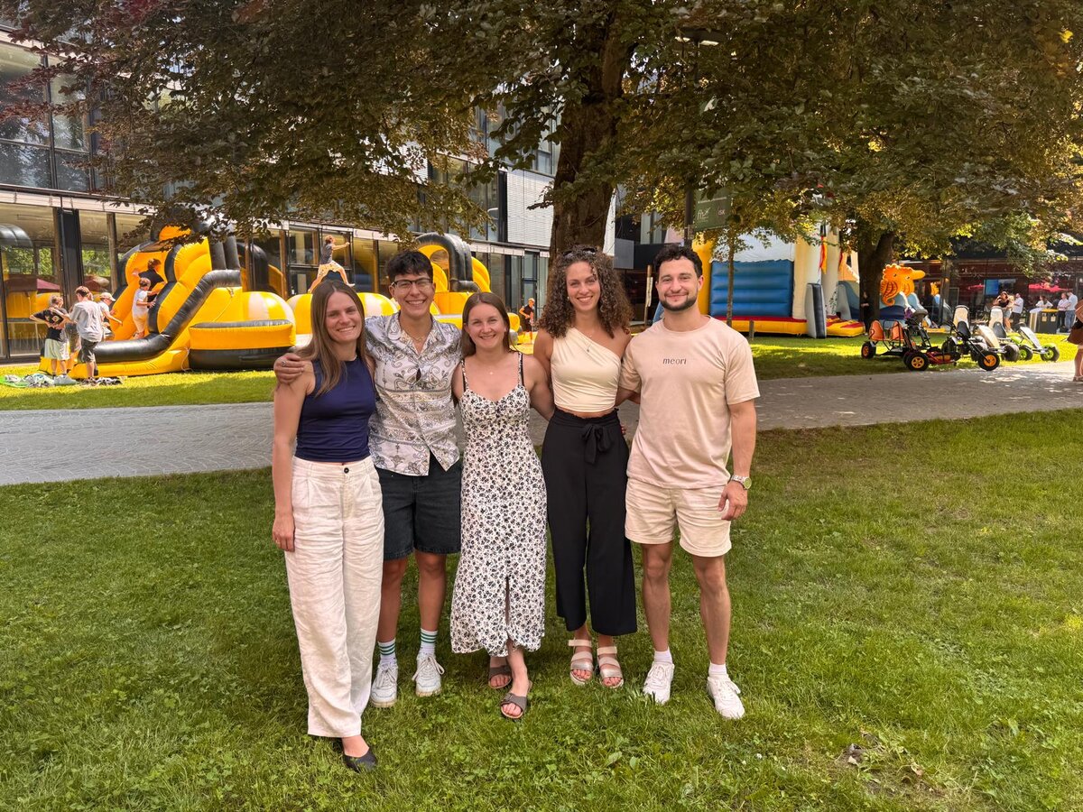 Fünf junge Erwachsene stehen lächelnd auf einer Wiese am Campus Kufstein. Im Hintergrund sind Kinder auf Hüpfburgen und Kettcars zu sehen, was auf ein Sommerfest oder Event hinweist.