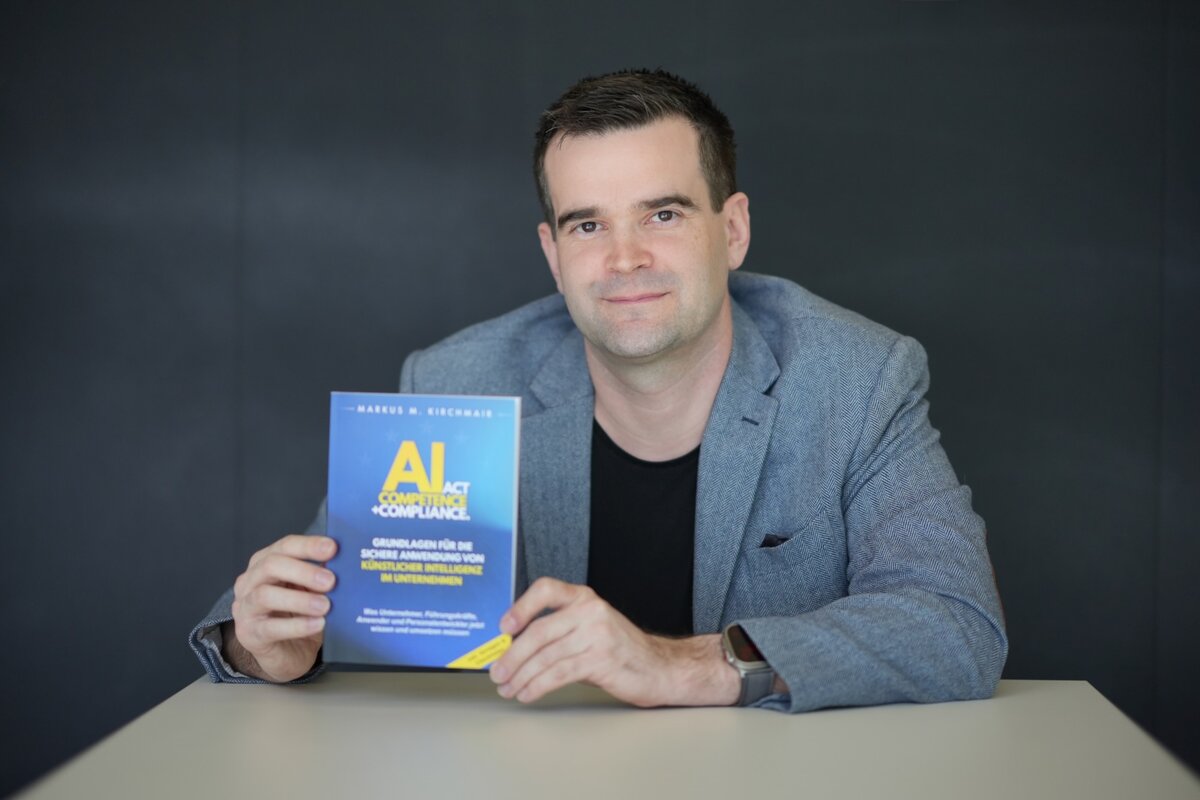 Mann sitzt vor einem dunkelgrauen Hintergrund am Tisch und hält ein Buch mit dem Cover in die Kamera.